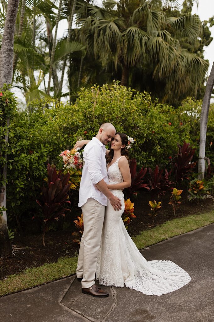 A bride and groom embrace one another and smile while standing on a path in front of lush green bushes and palm trees