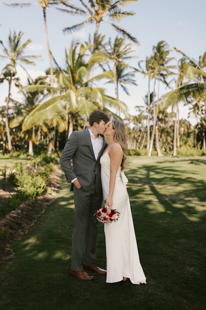 A bride and groom stand side by side and kiss one another while the bride holds her bouquet, behind them are palm trees