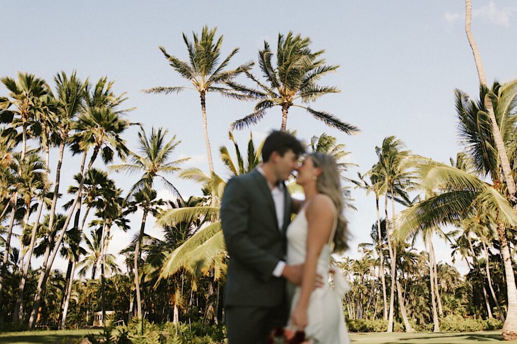 A bride and groom embrace in the foreground and are about to kiss during their elopement at Lanikūhonua on Oahu, behind them in focus are palm trees