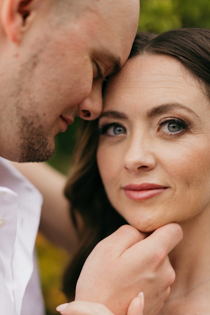 A bride smiles at the camera while the groom touches his forehead to hers with his eyes closed