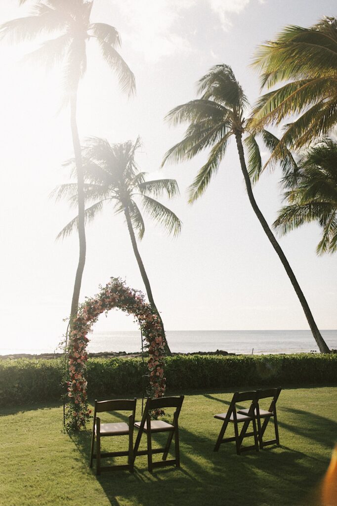 An elopement ceremony space set up with 4 chairs, a flower arch, and palm trees in front of the ocean
