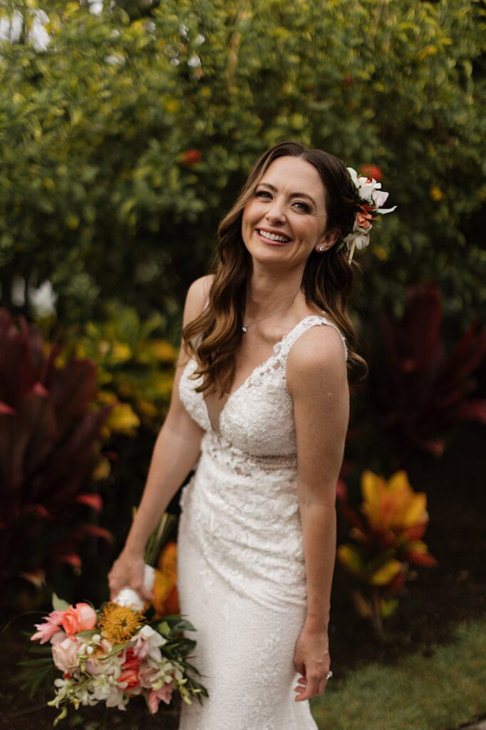 A bride holding a bouquet of flowers smiles at the camera in front of lush green bushes and large flowers