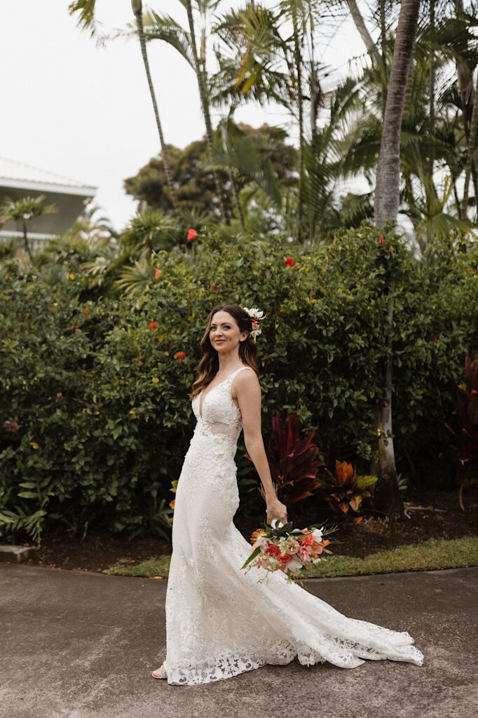A bride smiles at the camera with a bouquet in hand while walking along a path in front of large green bushes and palm trees