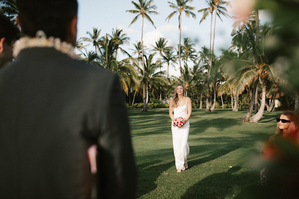 A bride smiles while walking towards the groom who has his back to the camera during their elopement ceremony at Lanikūhonua on Oahu, behind the bride are palm trees in a grass field