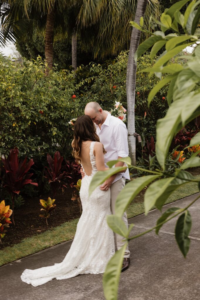 A bride and groom embrace one another and touch foreheads together while standing on a path lined with lush greenery and palm trees
