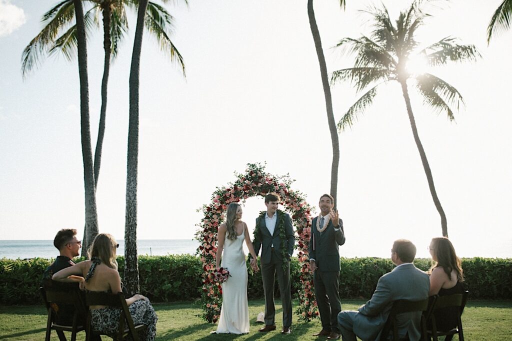 A bride and groom stand side by side and hold hands as their officiant speaks during their elopement ceremony at Lanikūhonua on Oahu with their flower arch, palm trees, and the ocean behind them, their parents are also seated watching