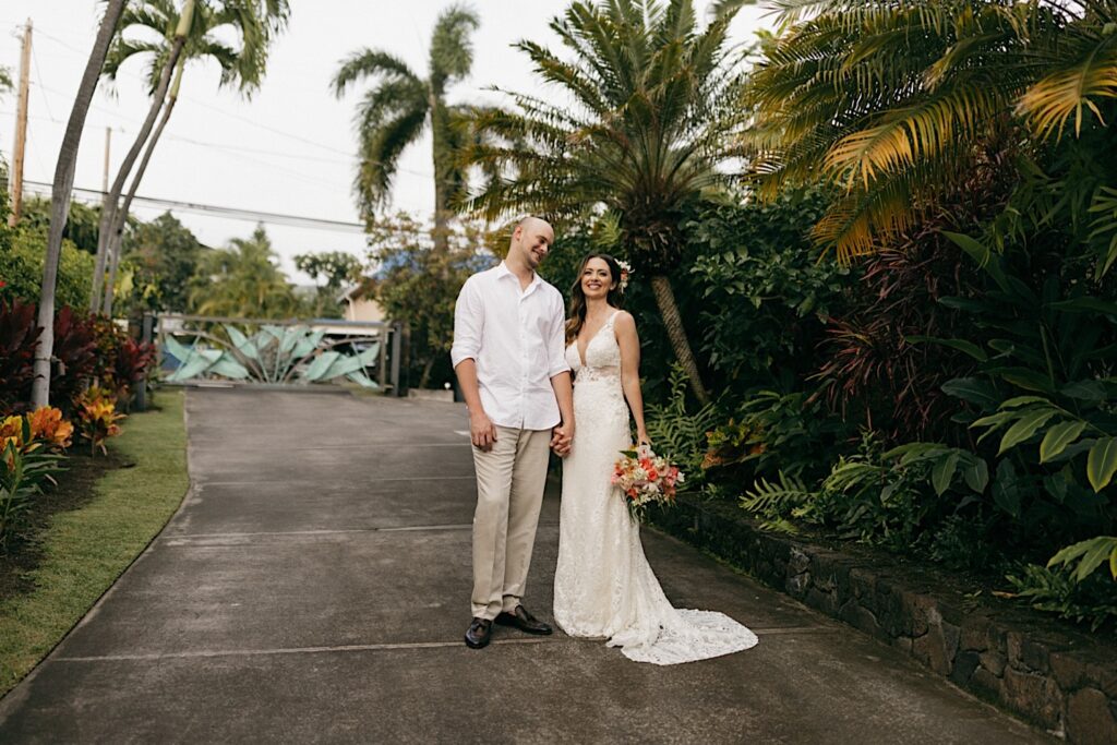 A bride and groom stand next to one another and hold hands while smiling at the camera on a path lined by pal trees and lush greenery before their intimate wedding on Hawaii's big island