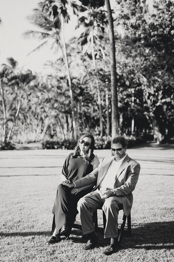 Black and white photo of a couple sitting in a grass field with palm trees behind them and holding hands while watching an elopement ceremony