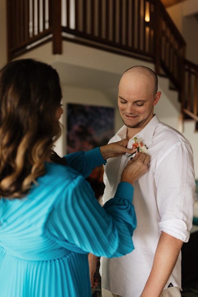 A groom smiles while looking down as his mother pins a flower to his shirt before his wedding