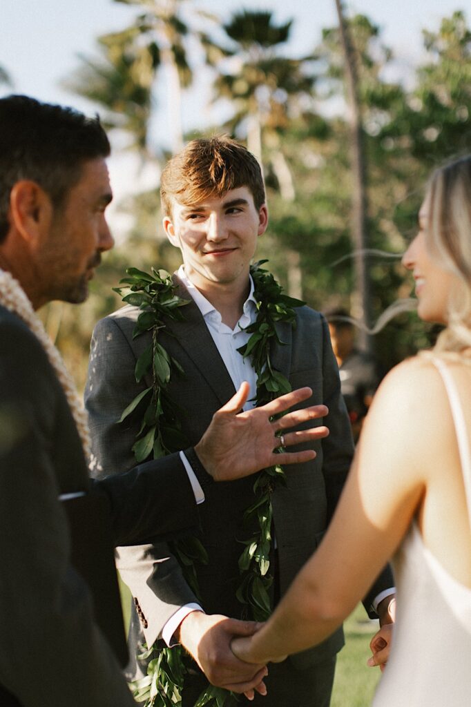 A groom smiles at the officiant of his ceremony while holding the bride's hands as she also smiles at the officiant