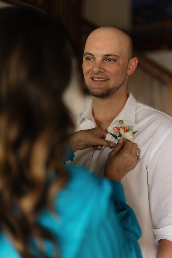 A groom smiles towards something off camera as his mother pins flowers to his shirt before his wedding