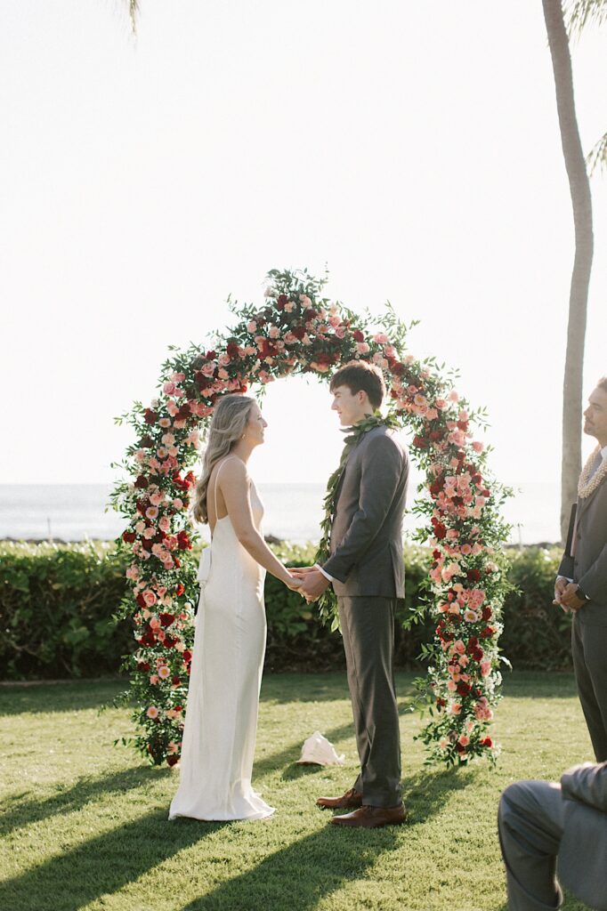 A bride and groom face one another and hold hands while standing in front of a flower arch and the ocean during their elopement ceremony