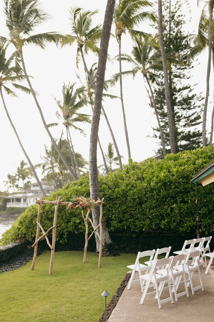 A ceremony space set up with white chairs, and a wooden archway with flowers sits empty before a wedding day, behind the space are palm trees and the ocean