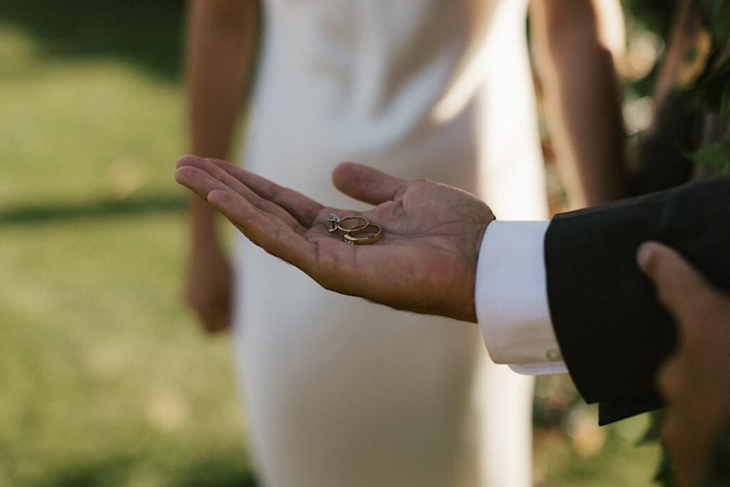 Close up photo of a hand holding two wedding rings with the bride in the background