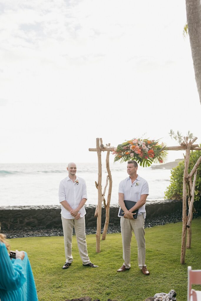 A groom stands with his officiant at his ceremony space in front of the ocean as the wedding ceremony begins