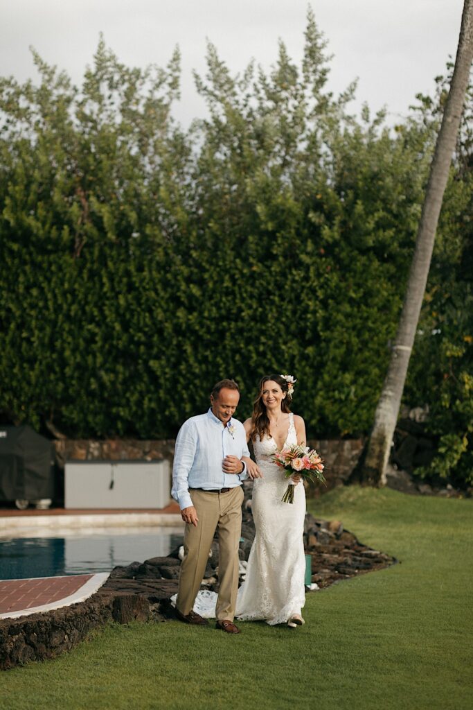 A bride smiles at the camera while walking alongside her father with their arms locked, behind them are a pool and a large wall of ferns