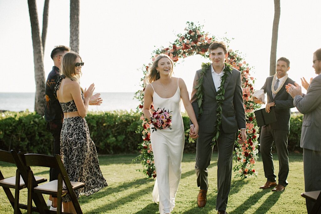 A bride and groom smile while holding hands and exiting their elopement ceremony at Lanikūhonua on Oahu, their officiant and parents clap for them as they make their exit with the ocean behind them