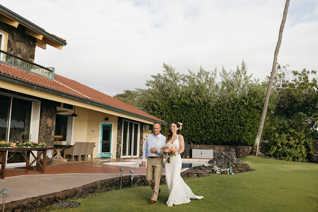 During an intimate wedding ceremony on Hawaii's big island, a bride smiles while walking alongside her father with their arms locked in front of a pool and their house