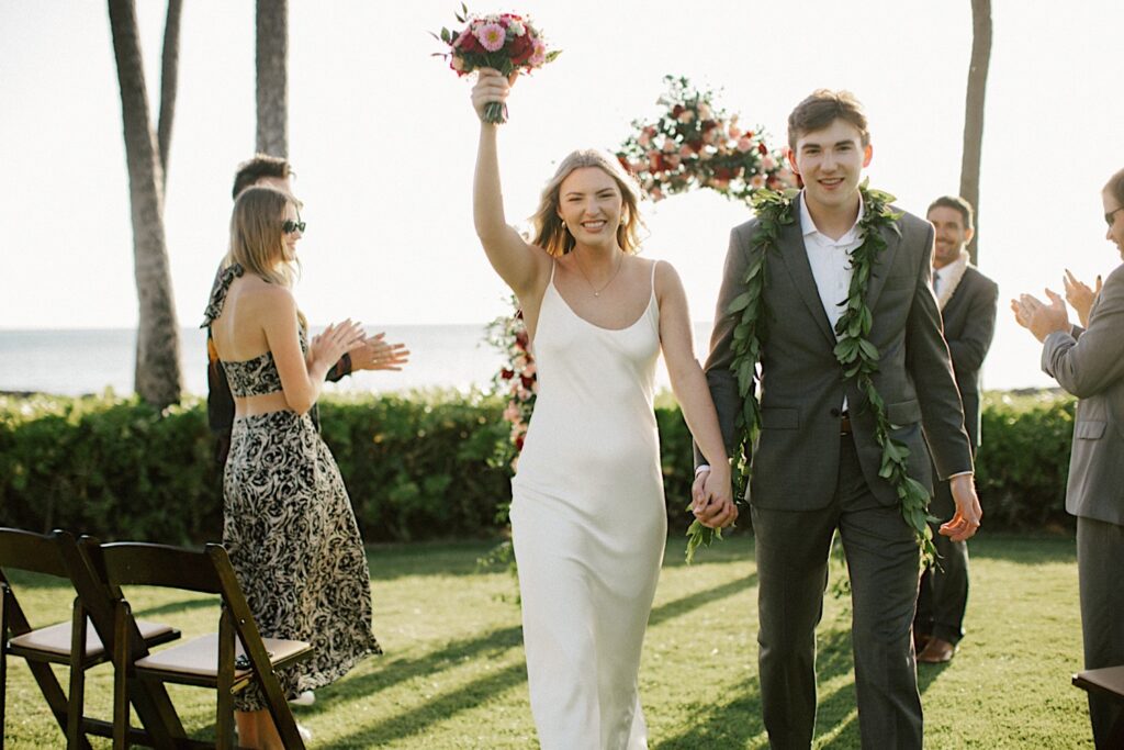 A bride and groom hold hands and smile at the camera as the bride lifts her bouquet in the air as they exit their elopement ceremony at Lanikūhonua on Oahu and their parents clap for them