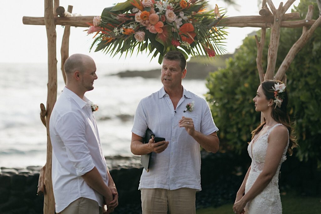 During an intimate wedding ceremony on Hawaii's big island with the ocean in the background, an officiant speaks while standing underneath a wooden archway in-between the bride and groom