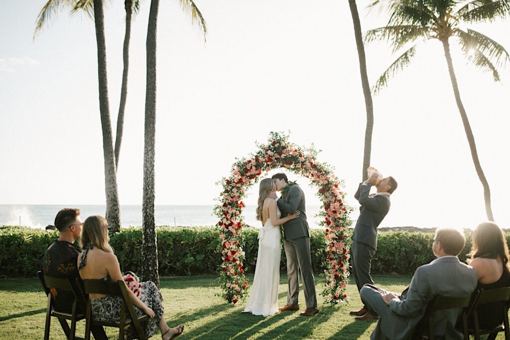 A bride and groom kiss under a floral archway as their officiant blows into a shell during their elopement ceremony at Lanikūhonua on Oahu