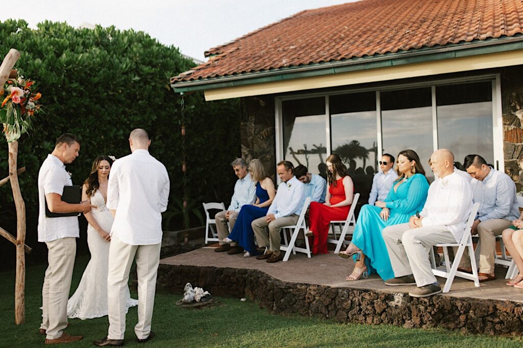 During an intimate backyard wedding on Hawaii's big island a bride and groom stand next to their officiant while their guests sit and watch
