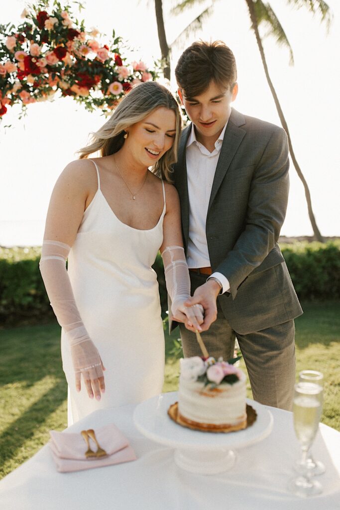A bride and groom cut their wedding cake together while smiling at it, behind them the sun is setting and a flower archway hangs overhead
