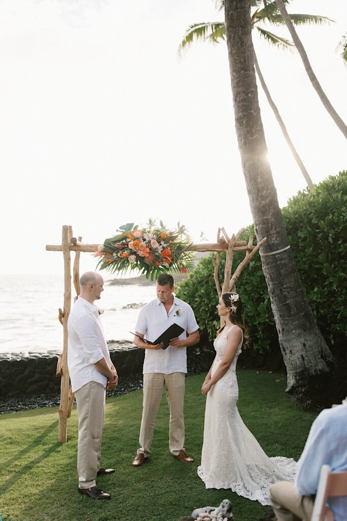 An officiant speaks while standing between a bride and groom during their wedding ceremony, behind them is the ocean and a wooden archway decorated with flowers