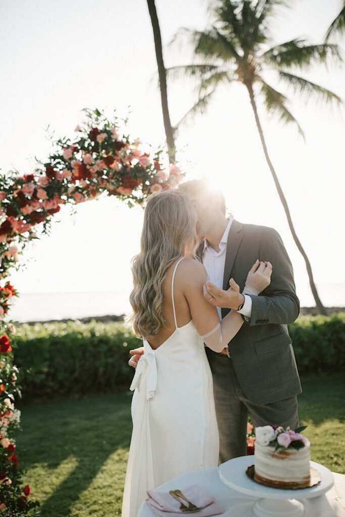 A bride and groom kiss one another after cutting their wedding cake, behind them is a flower archway and palm trees, the sun is also setting directly overhead