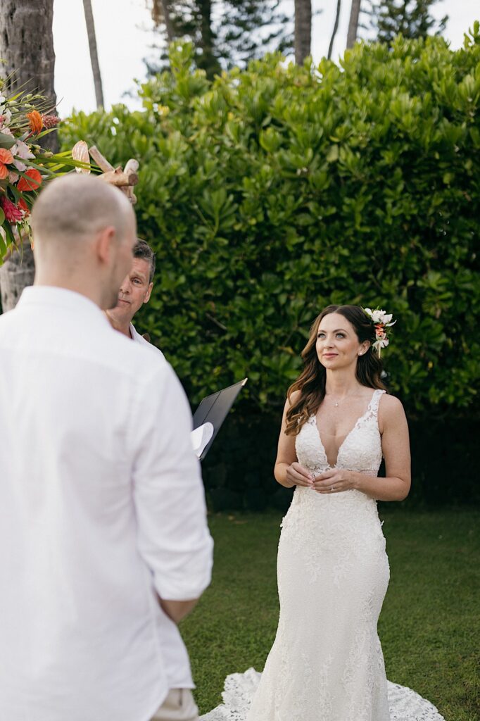 A bride smiles at a groom who is facing away from the camera as he reads his vows to the bride during their wedding ceremony