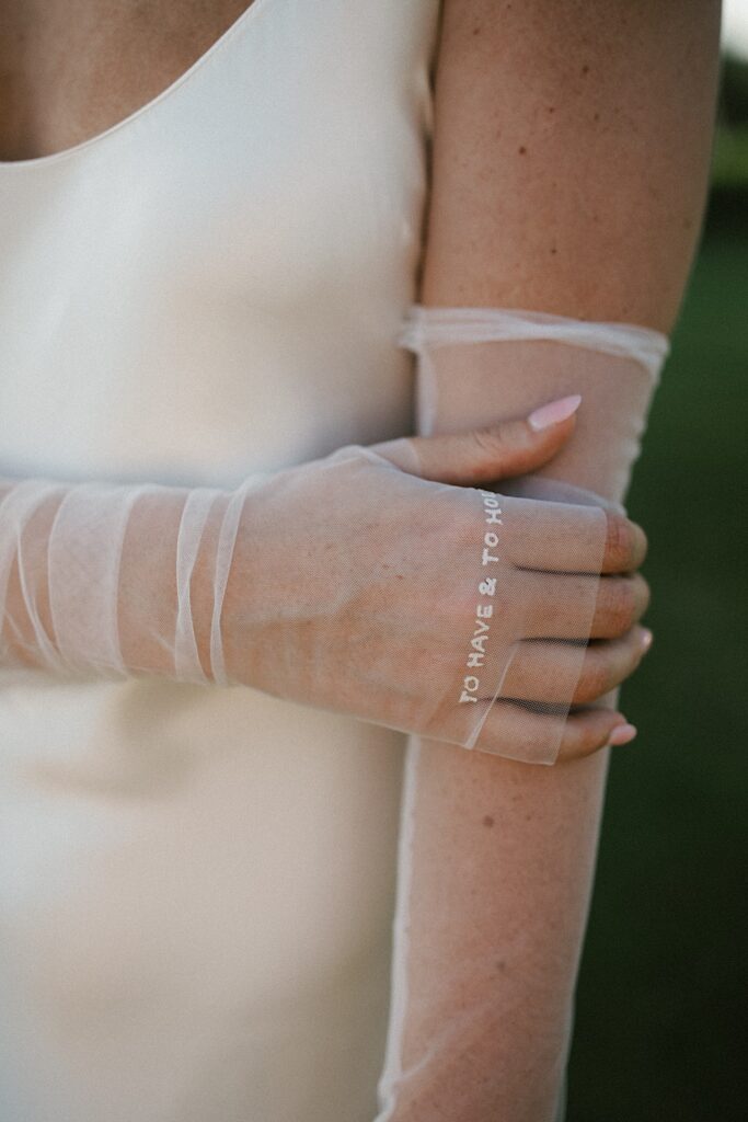Close up photo of sheer sleeves of a bride, she is holding her arm with one of her hands, on the sleeve are the words "to have & to hold"