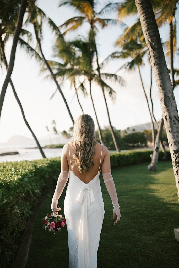 A bride with her back to the camera walks away along a bush line with palm trees around her and the ocean to the left