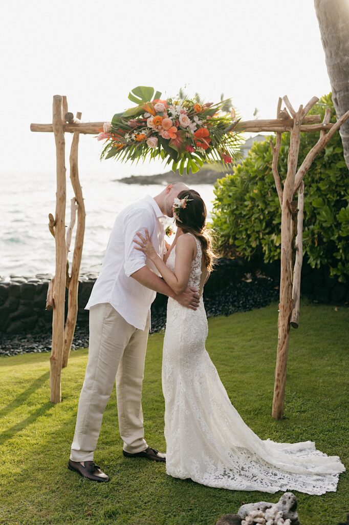A bride  and groom kiss one another during their wedding ceremony, behind them is a wood archway decorated with flowers and the ocean
