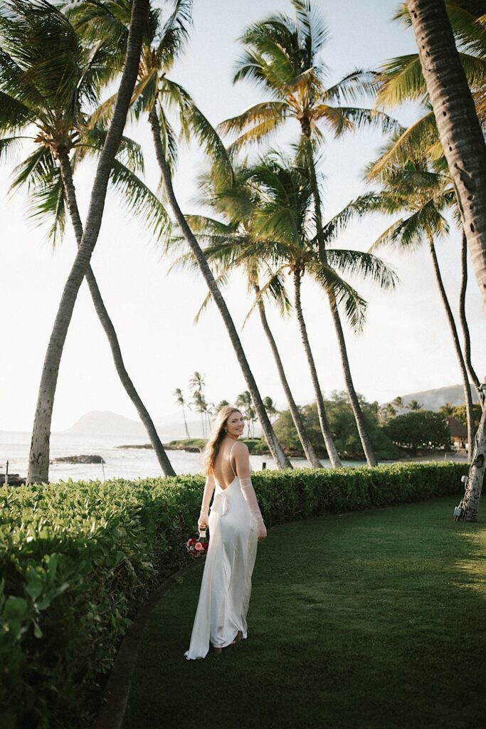 A bride with her back to the camera looks over her shoulder and smiles while standing next to a line of bushes, palm trees, and the ocean