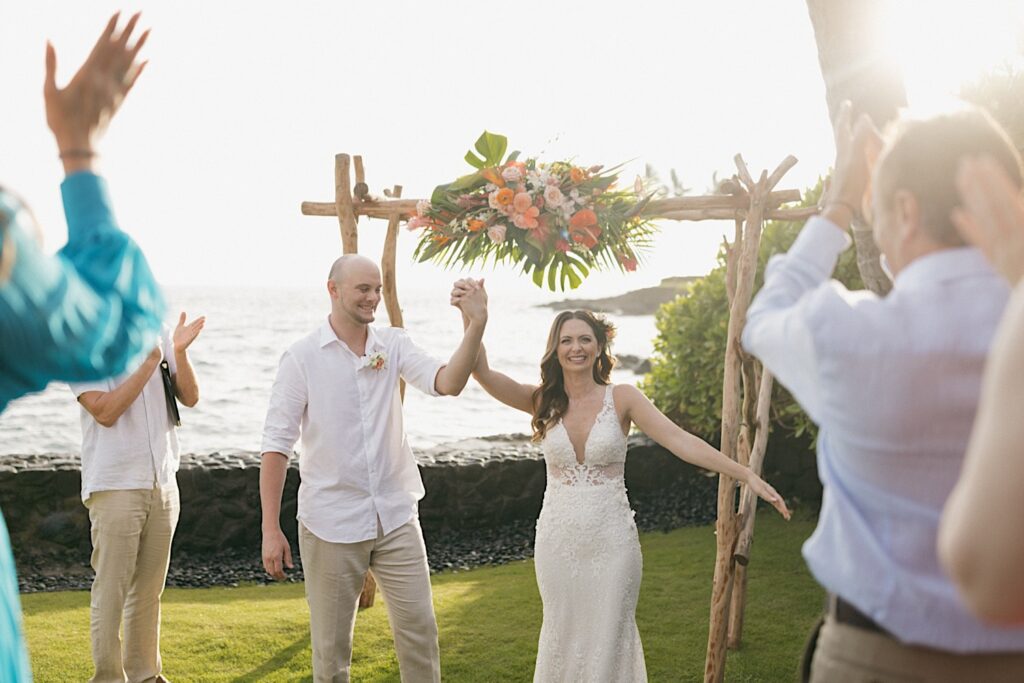 A bride and groom smile while holding hands and lifting them in the air towards the crowd who is cheering for them in the foreground of the photo during their intimate wedding ceremony on Hawaii's big island, behind them is the ocean and a wood archway decorated with flowers