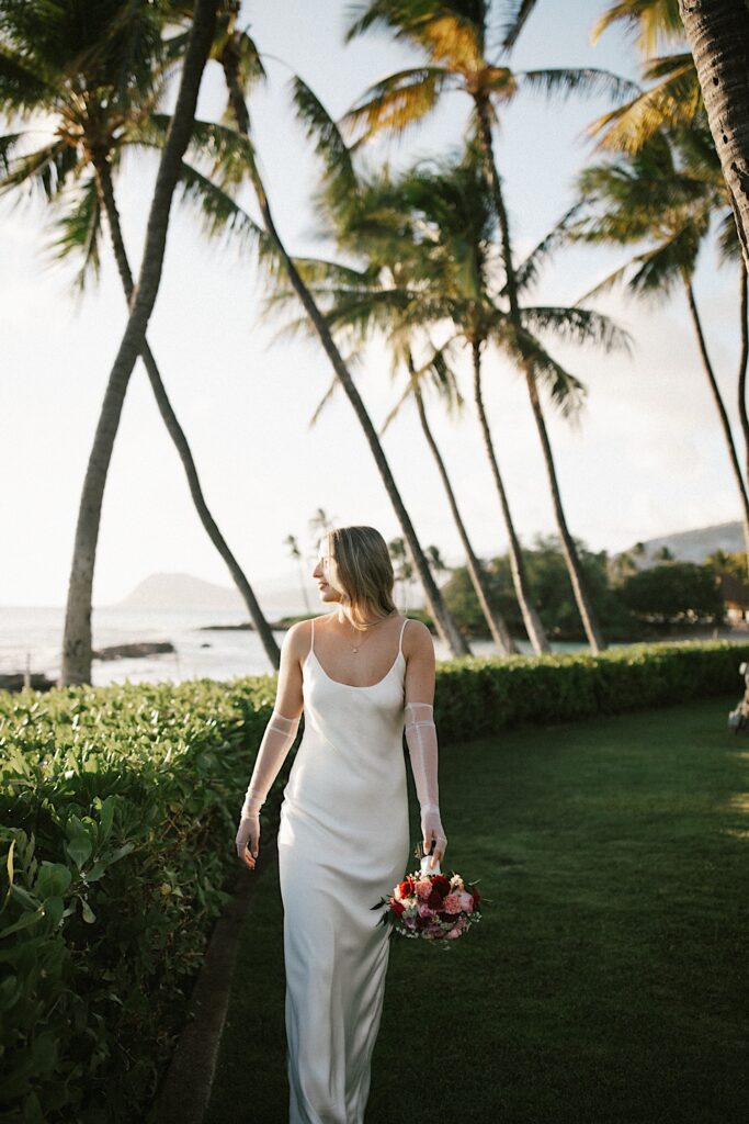 A bride walks towards the camera holding her bouquet and looks to the left towards the ocean as the sun sets, behind her is a line of bushes and palm trees