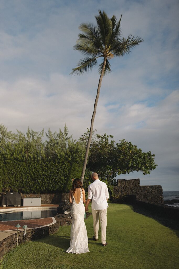 A bride and groom hold hands and walk away from the camera in a backyard towards a palm tree and a pool