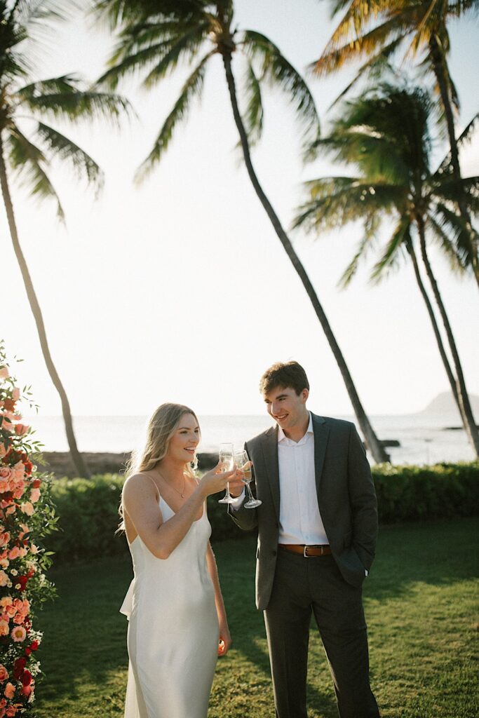 A bride and groom clink their champagne glasses together and smile as the sun sets behind them, in the background is the ocean and palm trees