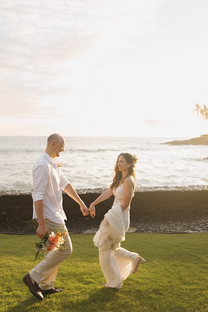 Standing in front of the ocean at sunset a bride and groom hold hands while facing one another and smiling, the bride is walking backwards while the groom walks towards her