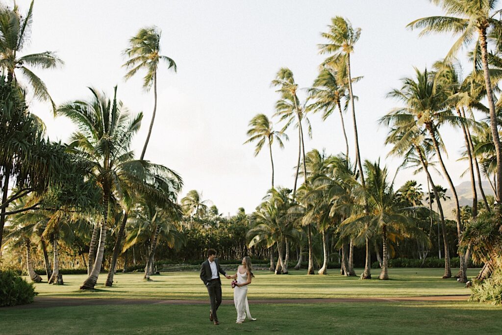 A bride and groom hold hands in the middle of a grass field surrounded by palm trees and smile at one another during their elopement at Lanikūhonua on Oahu