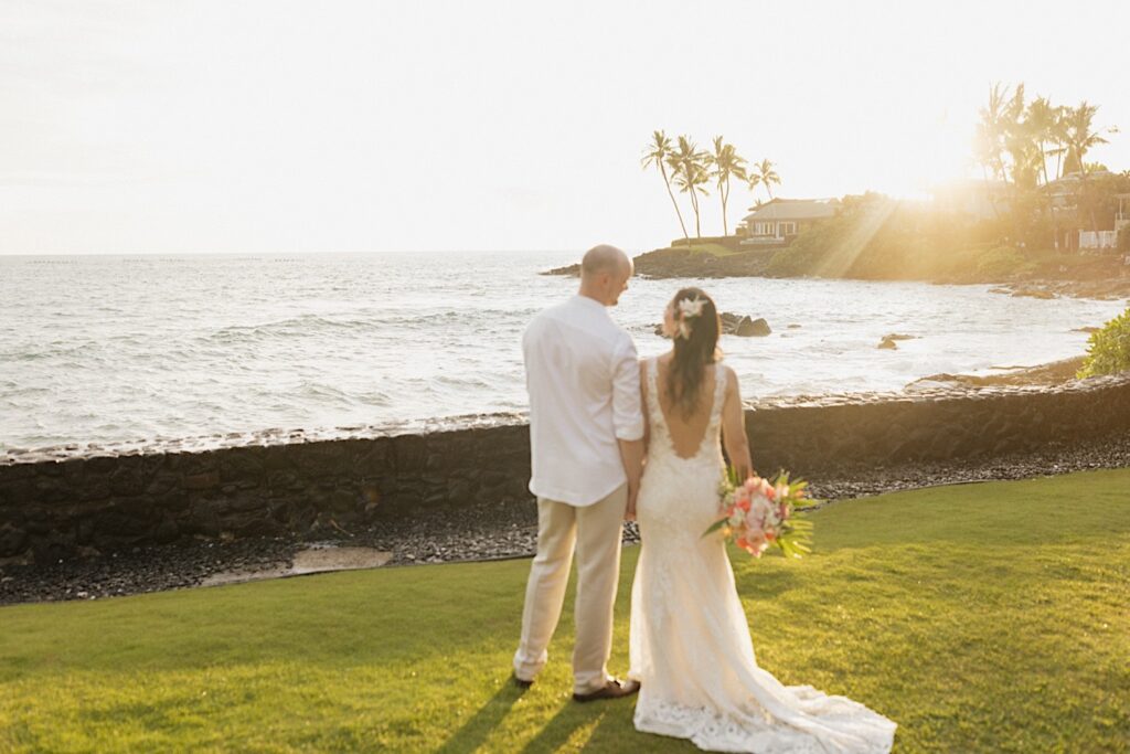 After their intimate wedding ceremony on Hawaii's big island a bride and groom hold hands and stand next to one another looking out at the ocean as the sun sets on it