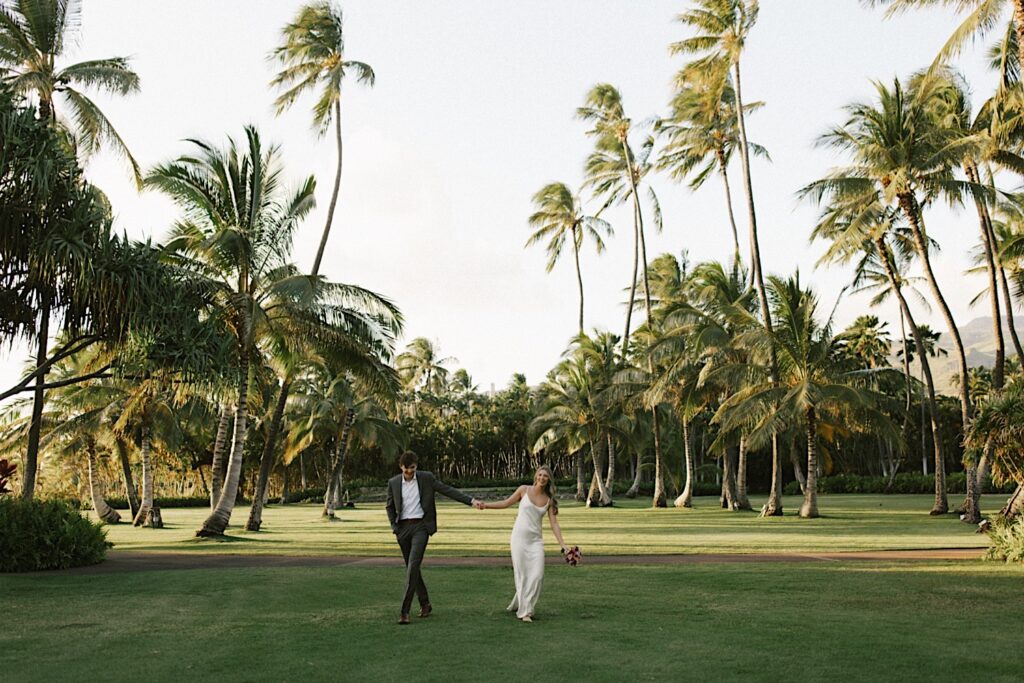 A bride and groom hold hands and walk towards the camera while smiling in the middle of a field surrounded by palm trees during their elopement at Lanikūhonua on Oahu