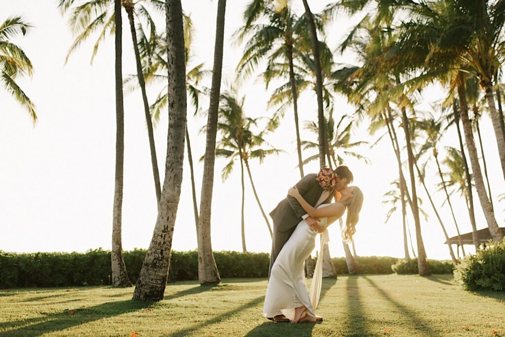 A bride is kissed and dipped by the groom while the two stand among palm trees as the sun sets during their elopement at Lanikūhonua on Oahu