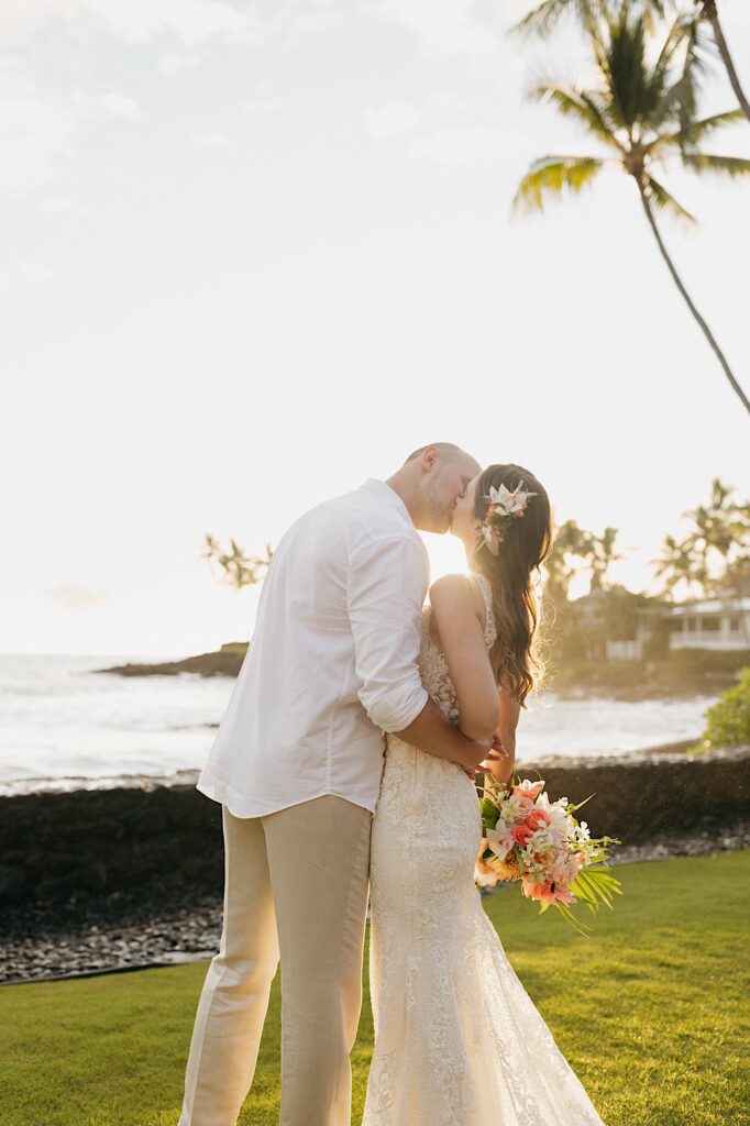During sunset a bride and groom kiss one another in a backyard with the ocean and palm trees behind them