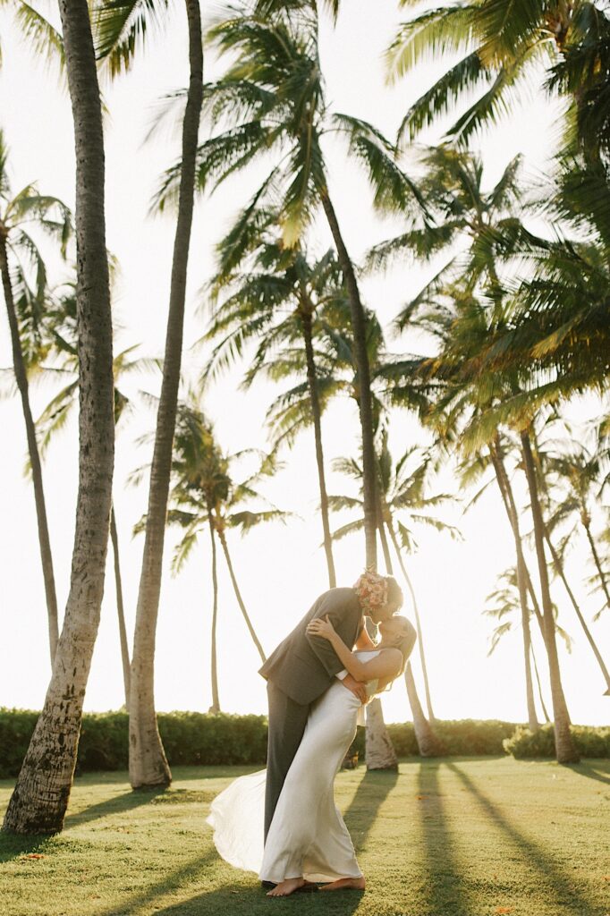 A bride is kissed and dipped by the groom during sunset while the two are surrounded by palm trees