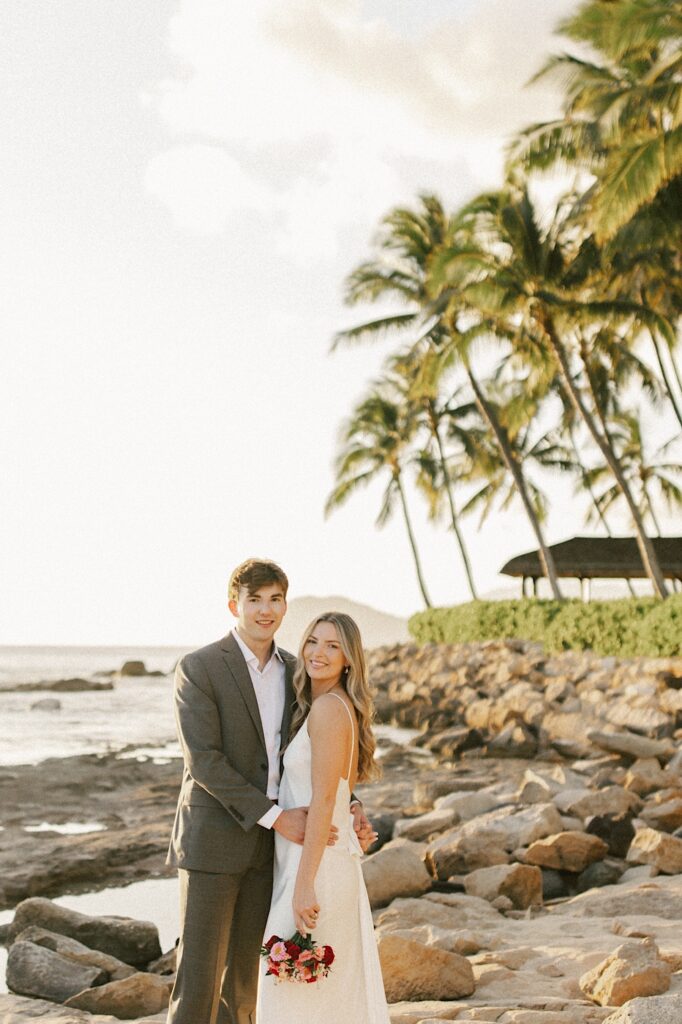 A bride is held by the groom as they both smile at the camera while standing on a rocky beach with palm trees and the ocean behind them at sunset