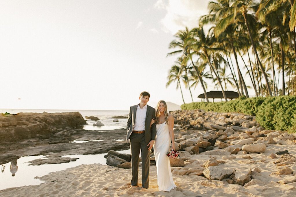 A bride and groom stand side by side and smile at the camera while the bride holds the groom's arm, they are on a rocky beach after their elopement at Lanikūhonua on Oahu with palm trees and the ocean behind them