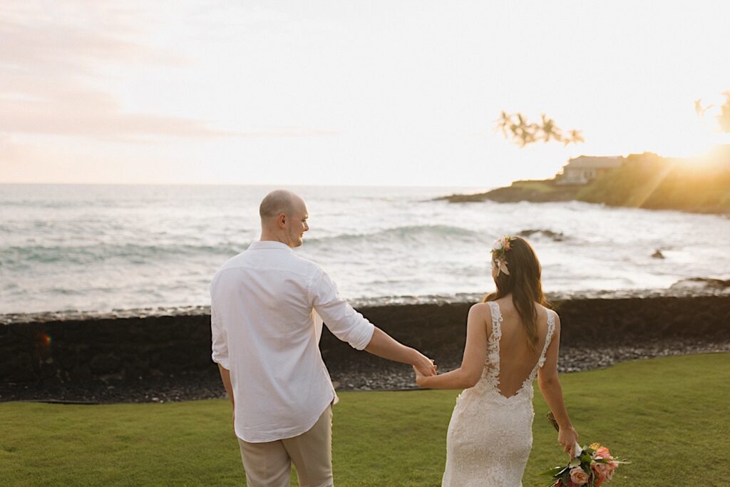 A bride and groom hold hands while looking out at the ocean after their intimate wedding ceremony on Hawaii's big island as the sun sets ocer the ocean