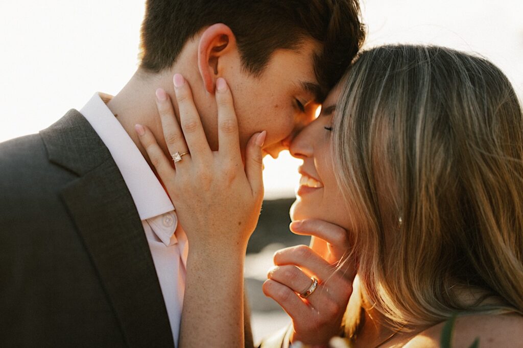 Close up photo of a bride and groom smiling as they are about to kiss one another with both of their wedding rings being shown off on their hands and the sun setting behind them