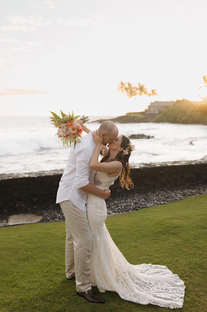 A bride and groom kiss one another while standing in a backyard in front of the ocean as the sun sets behind them